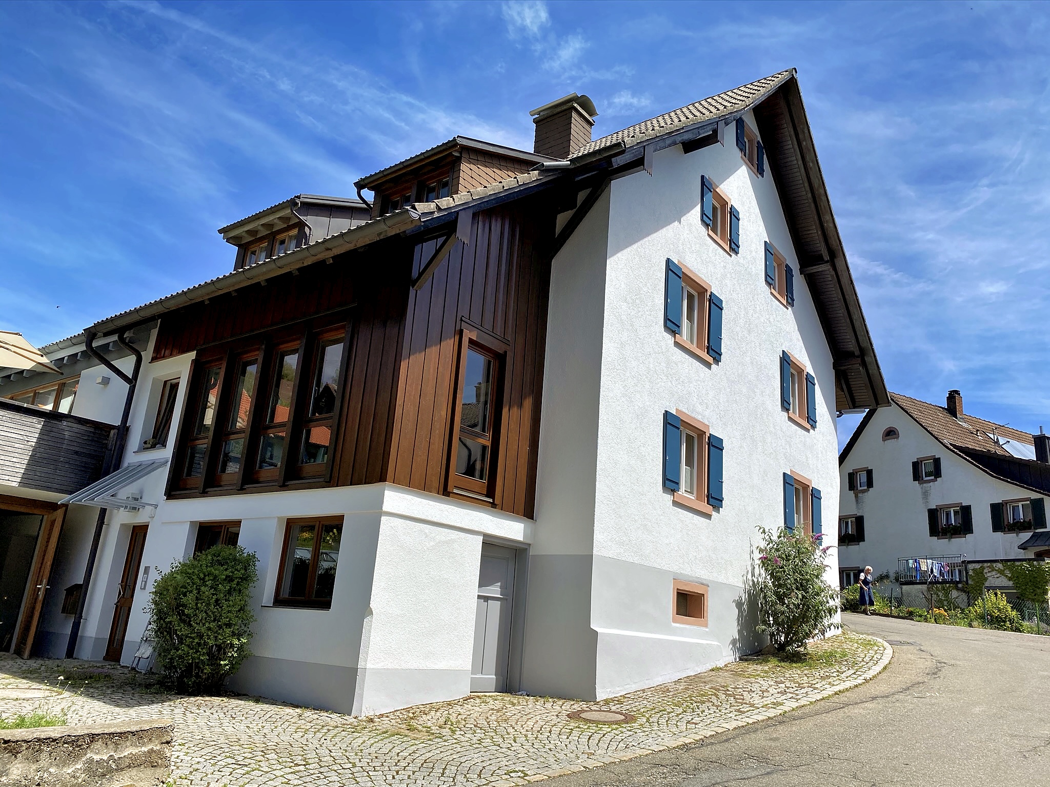 Ansicht eines Gebäude mit weißem Putz, Holzfassde und blauen Fenstern vor einem schönen blauen Himmel